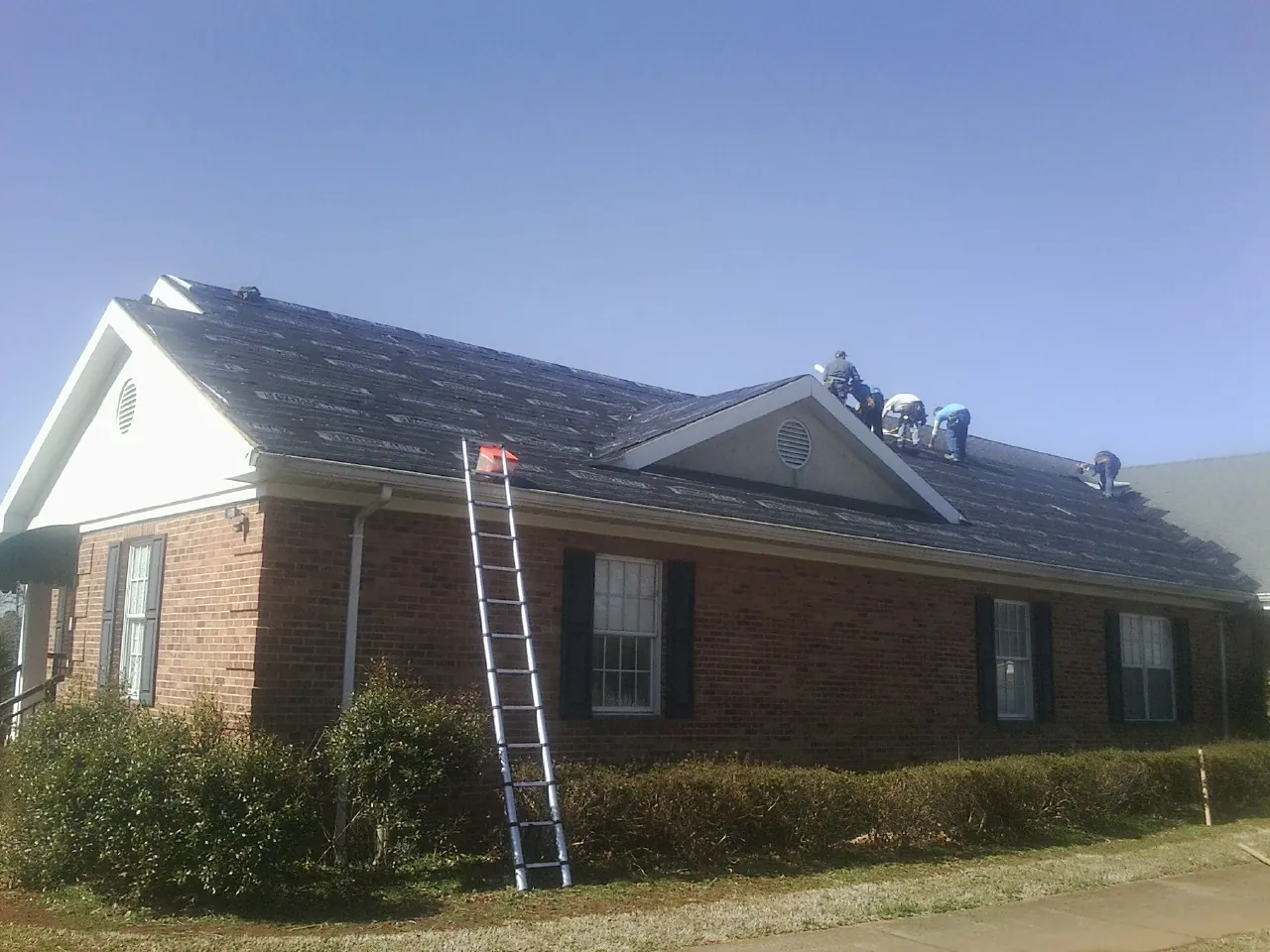 Crew tearing shingles off a brick ranch roof with a ladder set — Emergency Roof Repair work by our crew in Sandy Creek