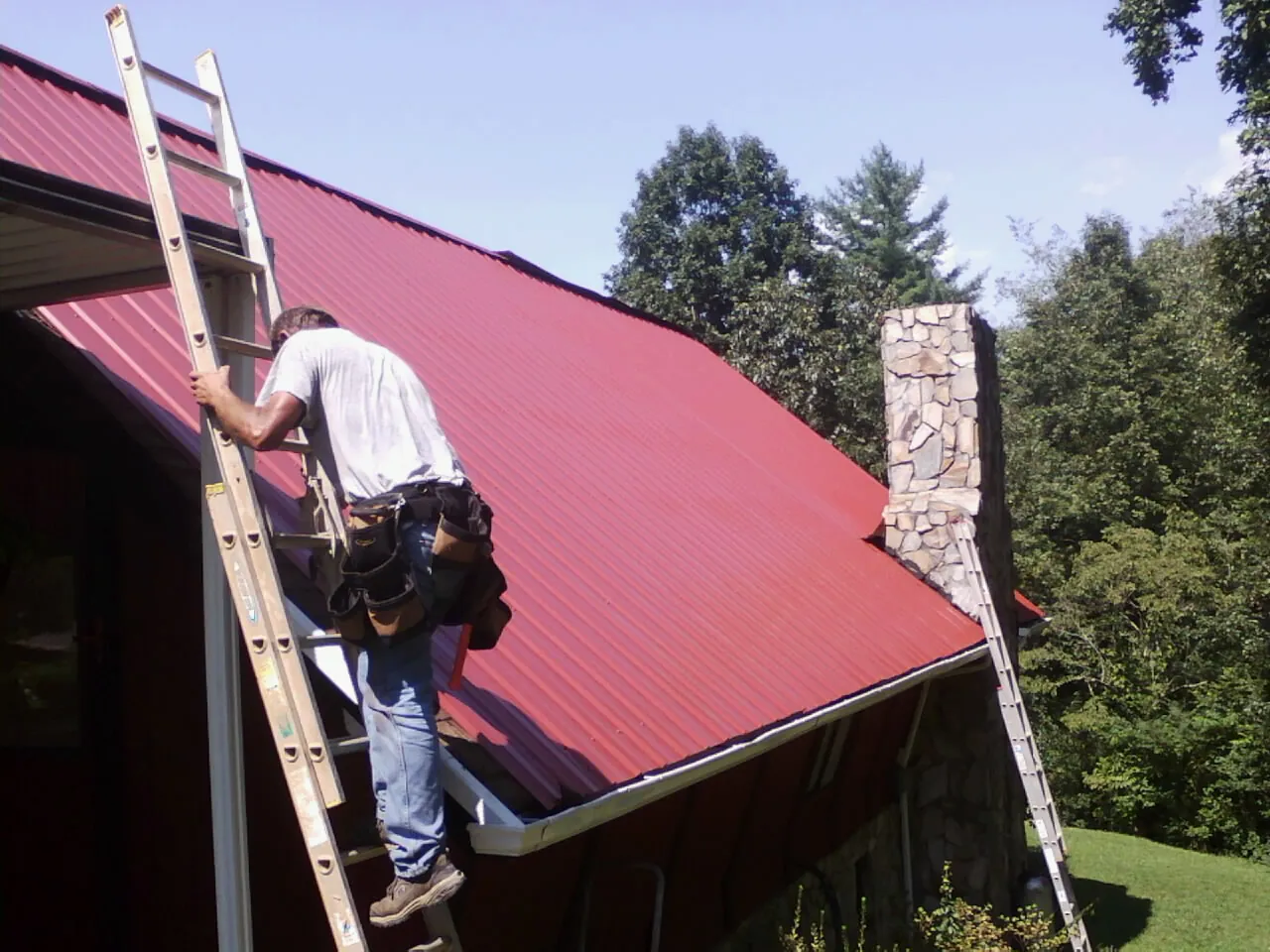 Roofer on a ladder beside a red metal roof and stone chimney — Soffit & Fascia Repair by Roofing Brothers in Sandy Creek